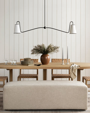 A minimalist dining room showcases a light wood table set with plates, glasses, and candles, a brown vase with greenery, woven chairs, a beige bench, and elegant lighting from the Makerspalm Jonas Linear Chandelier above. McGee & Co.