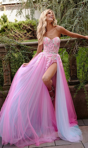A woman in the Jovani Sheer-Corset Prom Dress with floral embroidery, featuring pink and blue tulle and a high slit, smiles as she leans against an outdoor stone railing.