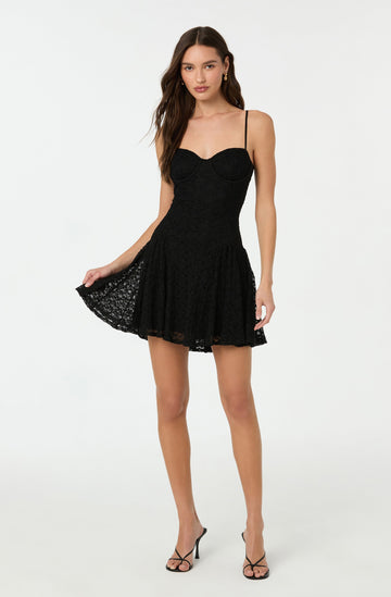 A young woman is wearing a black lace dress with a fitted bodice featuring a sweetheart neckline and thin spaghetti straps. The dress has a flared, above-the-knee skirt with a scalloped lace hem. She is also wearing black strappy high-heeled sandals.