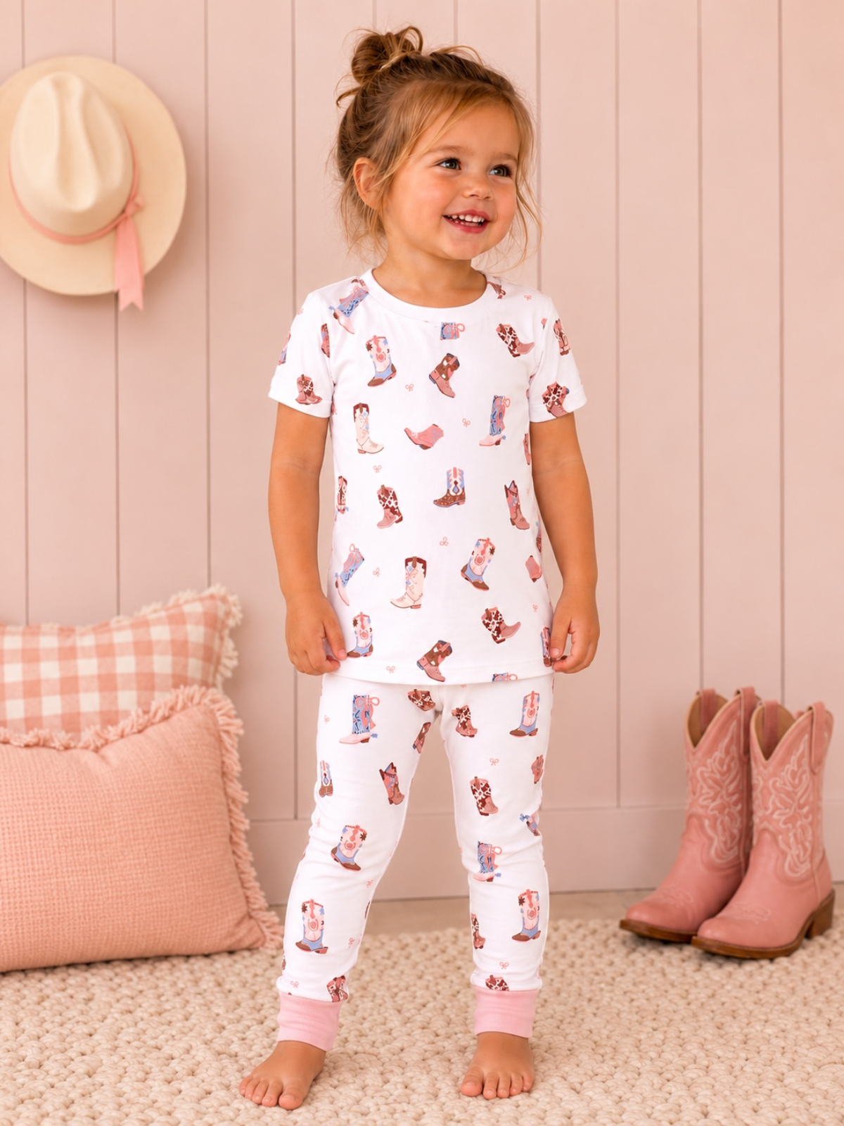 Smiling girl in patterned pajamas with boots design, standing on a rug near a pink pillow and boots.