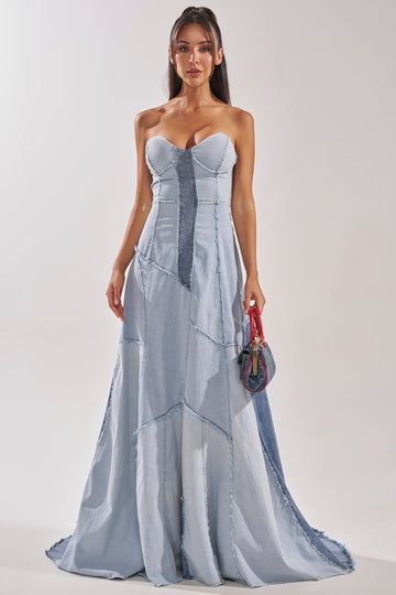 A woman wears the IT'S BRITNEY PATCHWORK DENIM MAXI DRESS, featuring strapless light and dark blue denim panels. She holds a small handbag, her long dark hair down, against a plain light-colored background.