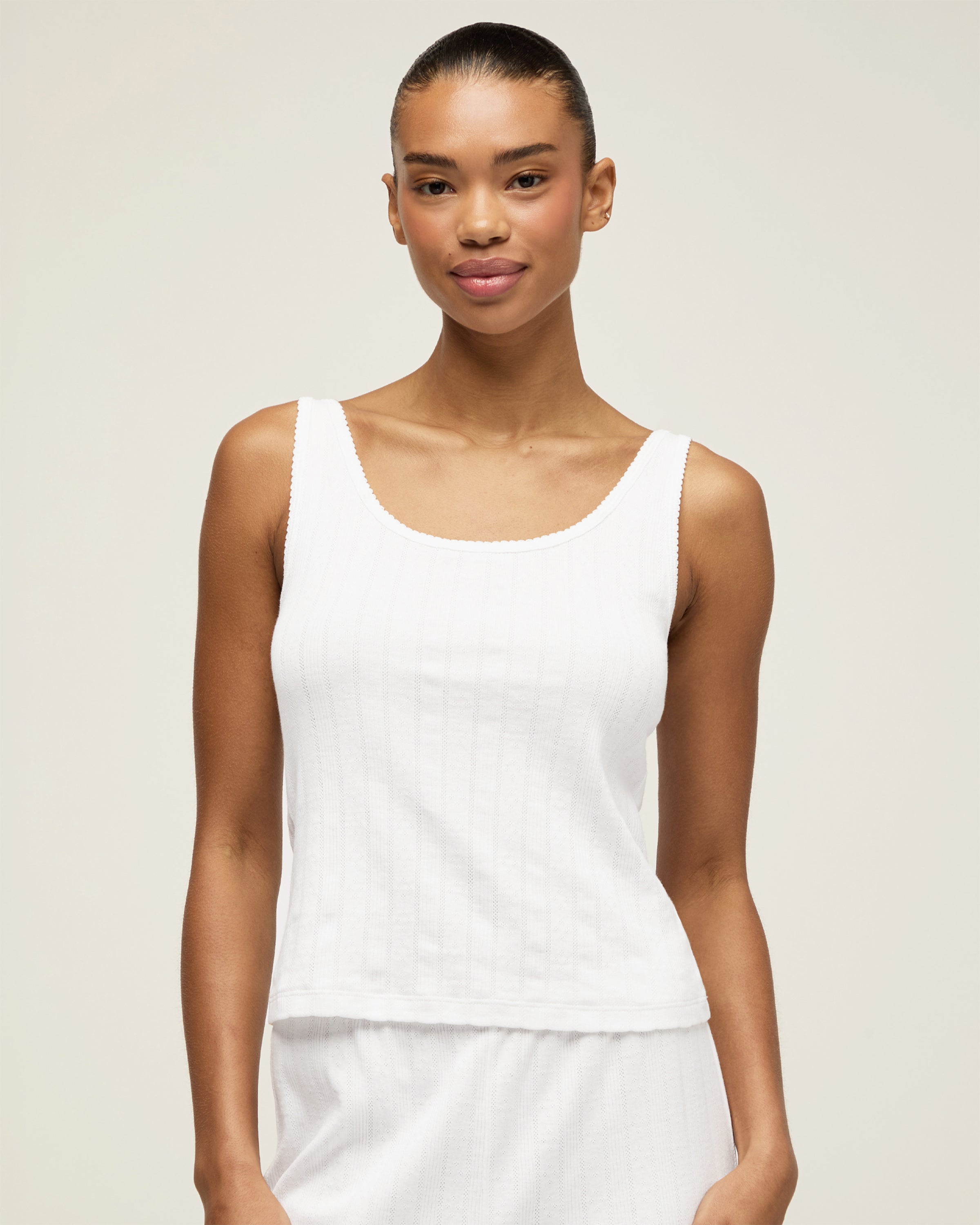 A woman with pulled-back hair wears the Women's Pointelle Tank in White and matching bottoms, standing against a plain light background and smiling softly at the camera.