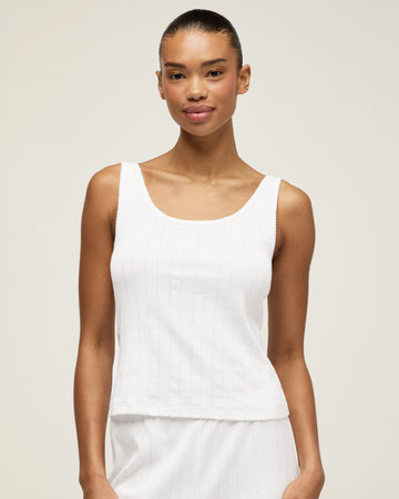 A woman with pulled-back hair wears the Women's Pointelle Tank in White and matching bottoms, standing against a plain light background and smiling softly at the camera.