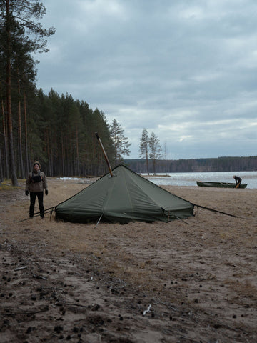 Person standing next to Savotta Hawu 8 tent on a lakeside beach with trees and water in the background.