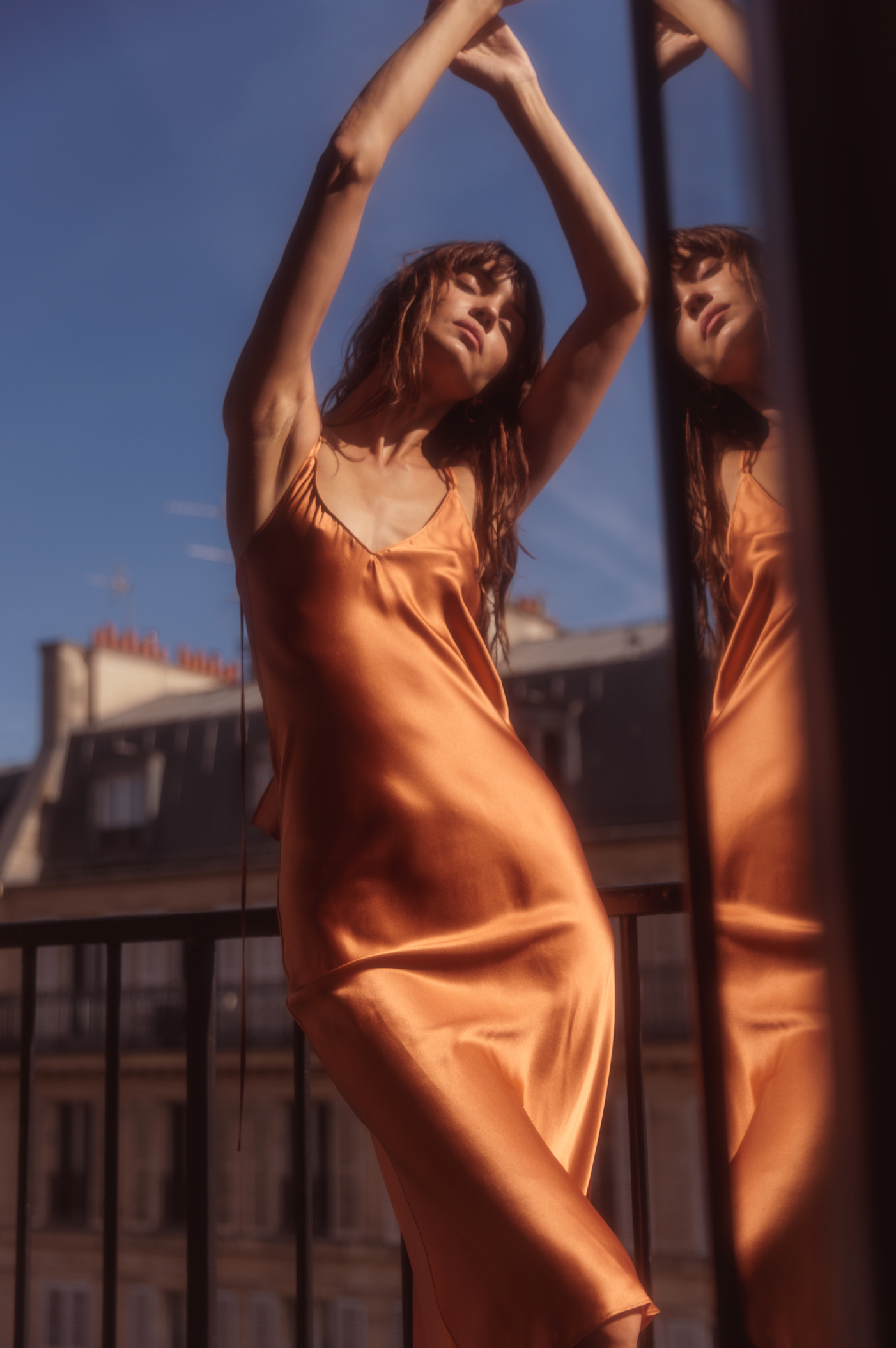 A woman posing with her arms raised, creating an elegant and fluid silhouette as she wears a Catherine Gee Emma Bronze Slip Dress. The dress is silky, bronze-colored, features thin straps, and has a V-neckline. In the background, a cityscape with buildings and a clear blue sky enhances the sophisticated urban vibe, while reflections in the glass add depth and visual interest to the composition.