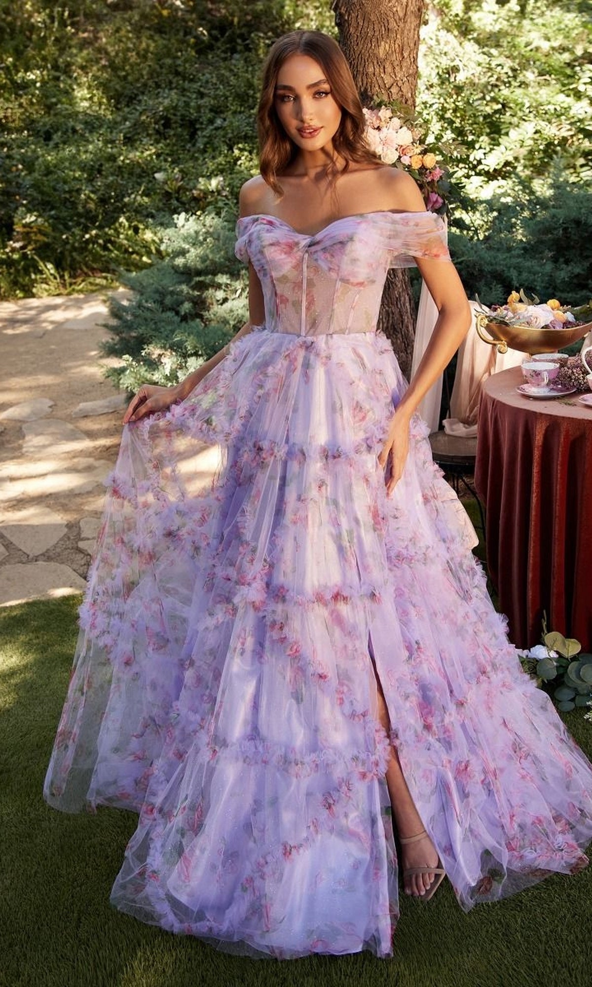 A woman wearing the Andrea & Leo A1286 long purple print prom dress stands outdoors near a tea table, surrounded by lush greenery.