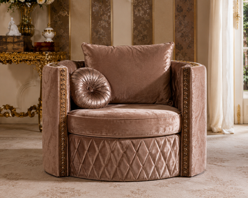 A luxurious upholstered armchair in a warm taupe velvet fabric, featuring a diamond-quilted base, flared arms with gold trim, a matching back cushion, and a round tufted accent pillow, set in an elegant room with gold-trimmed furniture and cream drapery.