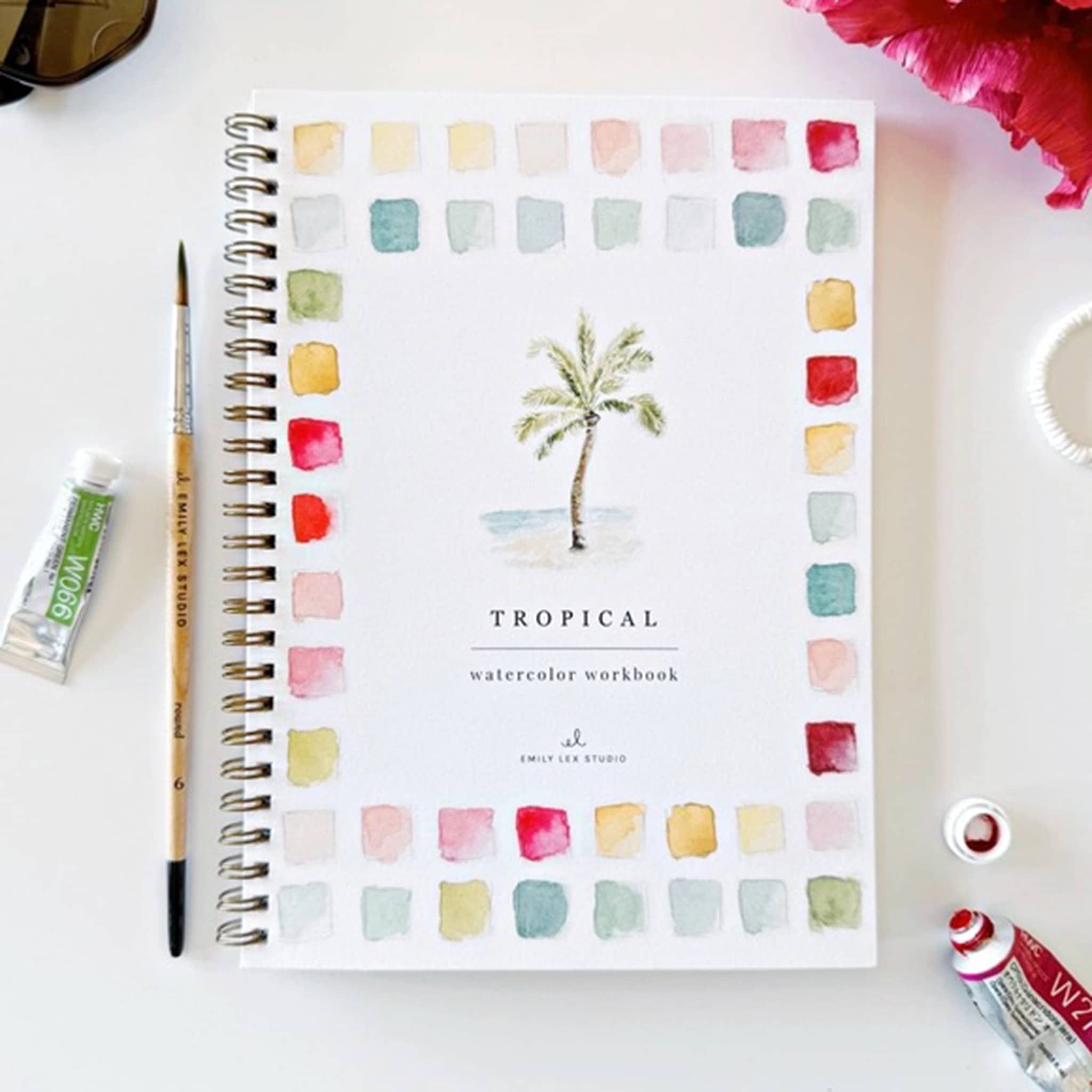 Front cover of the Tropical Watercolor Workbook with a palm tree design, on a white surface with a paintbrush, tubes of paint, and a flower.