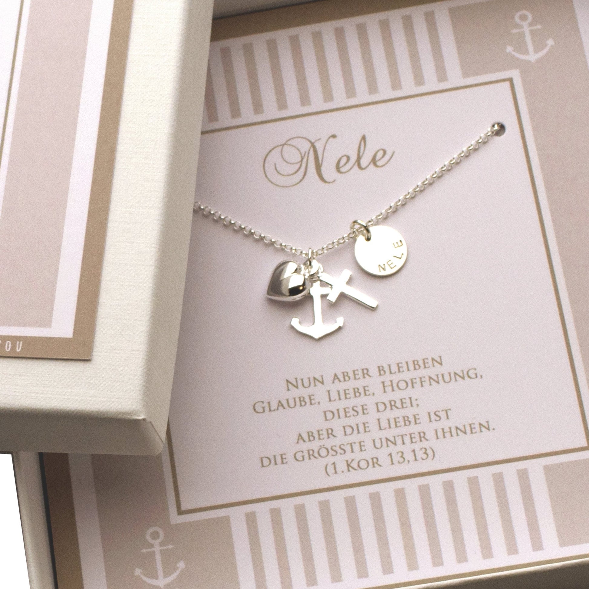Personalisierte Halskette aus 925 Sterling Silber mit Anhängern für Glaube (Kreuz), Liebe (Herz) und Hoffnung (Anker) sowie einem Gravurplättchen mit dem Namen „Nele". Die Kette liegt in einer Geschenkbox auf einer Karte mit dem Spruch aus 1. Korinther 13,13. Handgemacht von Bloomgart.