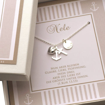 Personalisierte Halskette aus 925 Sterling Silber mit Anhängern für Glaube (Kreuz), Liebe (Herz) und Hoffnung (Anker) sowie einem Gravurplättchen mit dem Namen „Nele". Die Kette liegt in einer Geschenkbox auf einer Karte mit dem Spruch aus 1. Korinther 13,13. Handgemacht von Bloomgart.