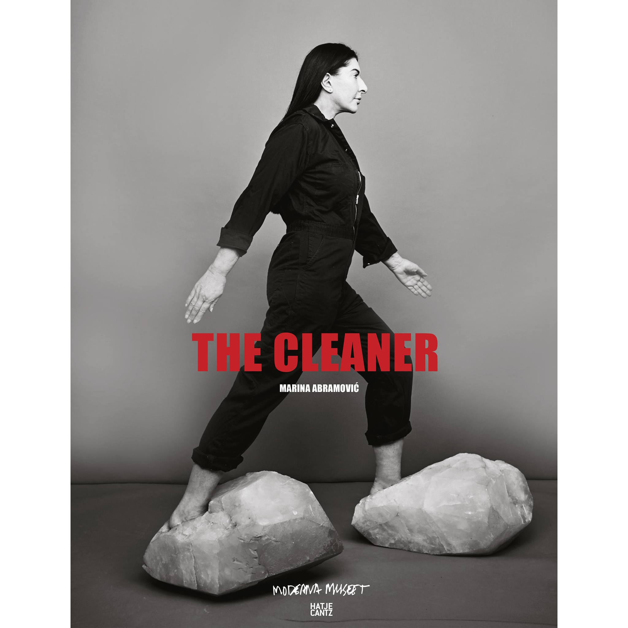 Eine Schwarz-Weiß-Fotografie von Marina Abramović, die in einem dunklen Overall barfuß über zwei große, helle Kristalle geht. In roten Großbuchstaben steht der Titel THE CLEANER über dem Bild.