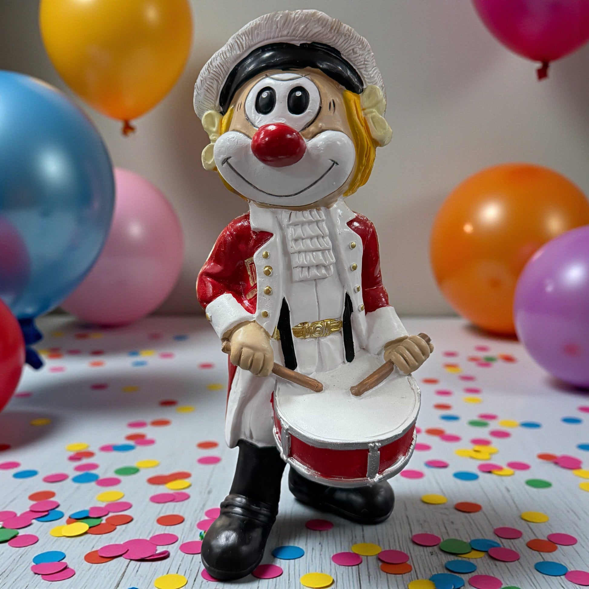Große Clown-Dekofigur mit roter Nase in rot-weißer Trommler-Uniform, ca. 20 cm groß, stehend dargestellt und eine Trommel vor dem Körper haltend, auf einer Tischfläche mit Konfetti, im Hintergrund bunte Luftballons.